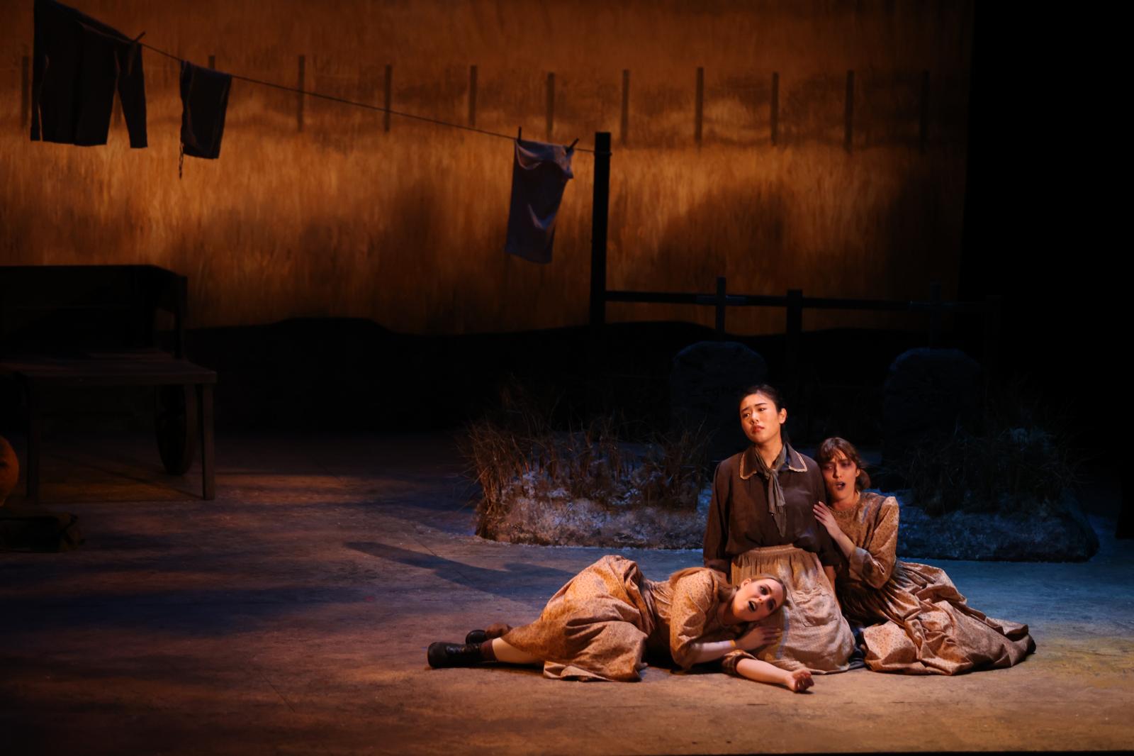 3 women clad in pioneer costumes sit on stage. A central figure appears somber but tall on stage, while another woman leans into her shoulder, and the third lays her head in the strong woman's lap. 