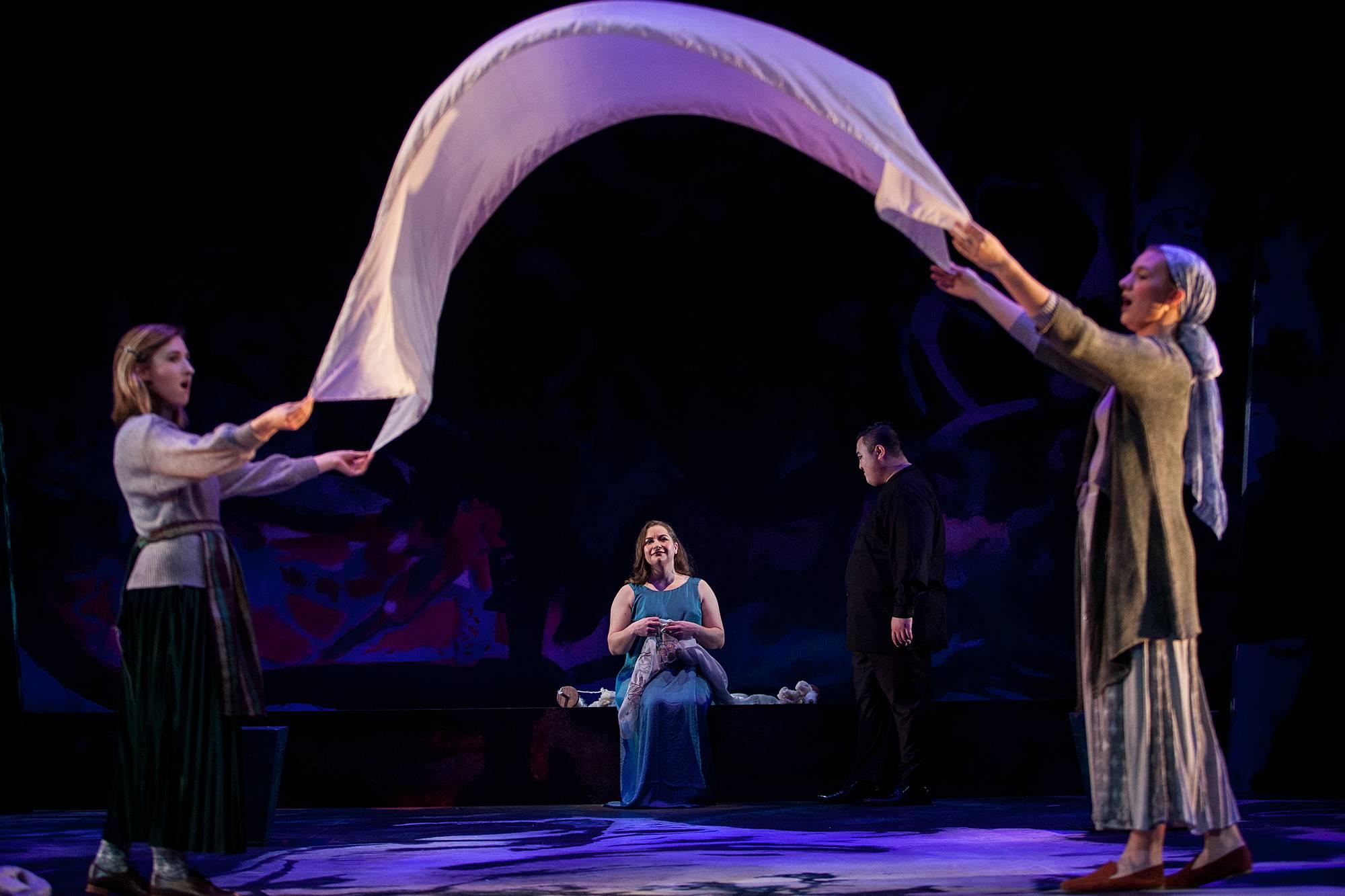 two women hold a billowy piece of white fabric up in a way that produces a cloth arch. Behind in the arch in the center, a woman in a Grecian dress stands alone.