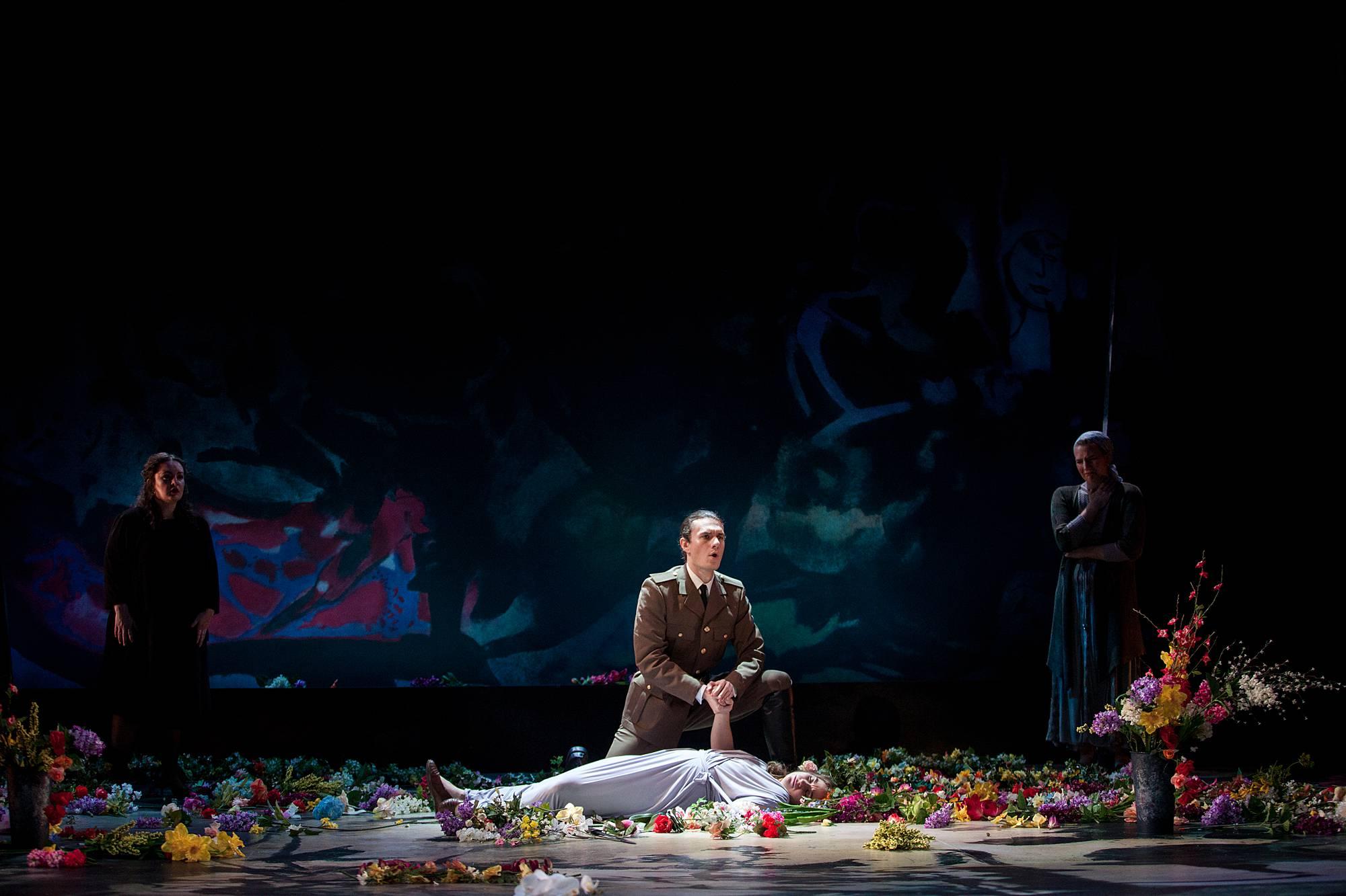 a man kneels on stage holding the hand of a dead woman, who is surrounded by flowers, as two mourners look on in the background. 