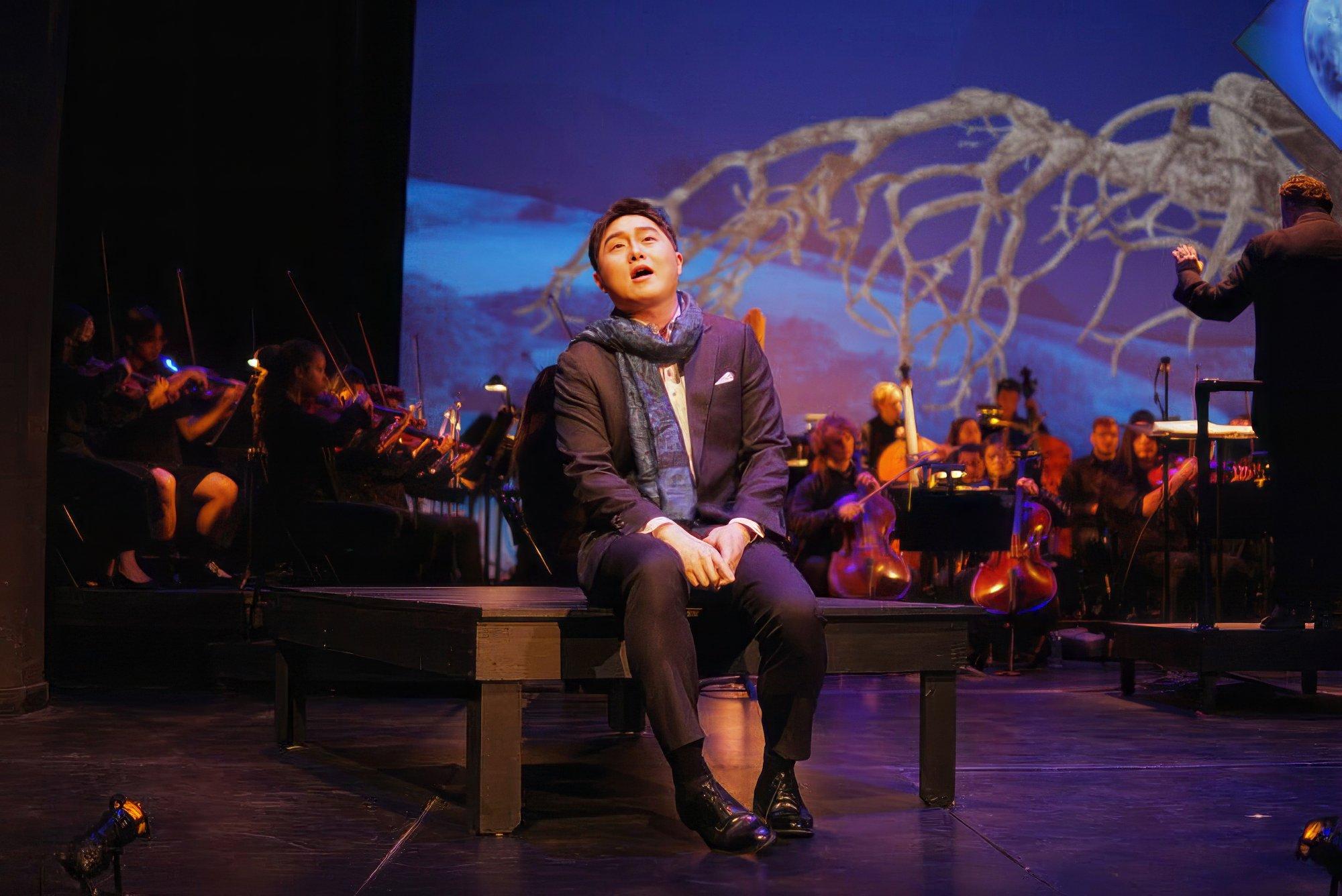 production still from the gala: A singer sits on a bench in the foreground dressed in a suit and scarf, with an orchestra on stage behind him.