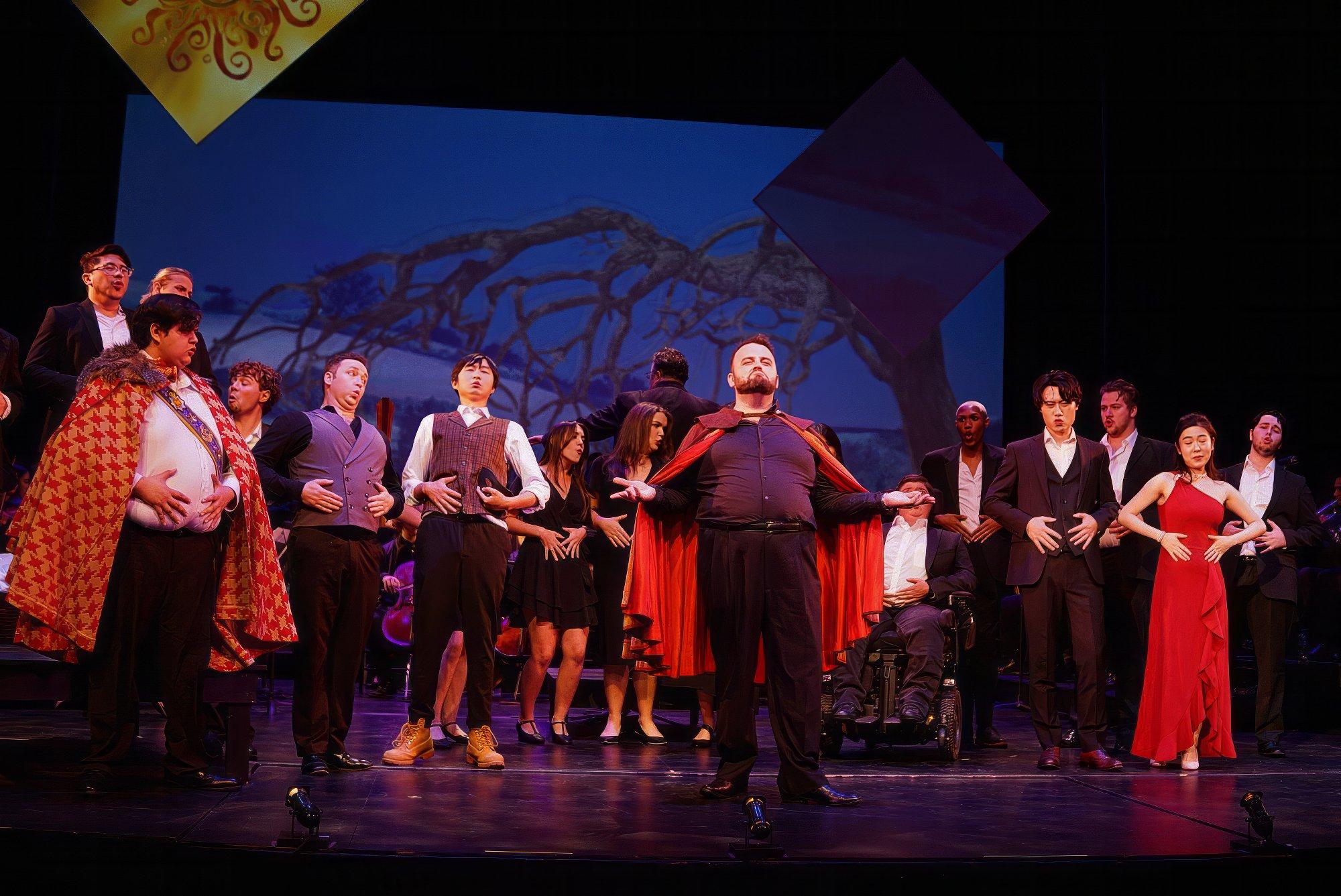 production still a group of singers on stage, in formal dress. a singer stands in front, arms outstretched, wearing a red cape. 