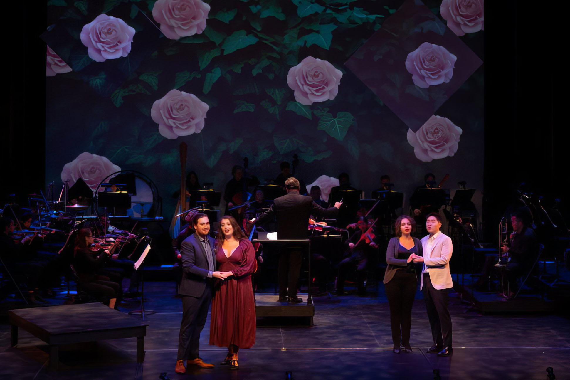 Production still: two embracing couples stand down stage with an orchestra behind them. The background is a projection of a pink rosebush in full bloom.