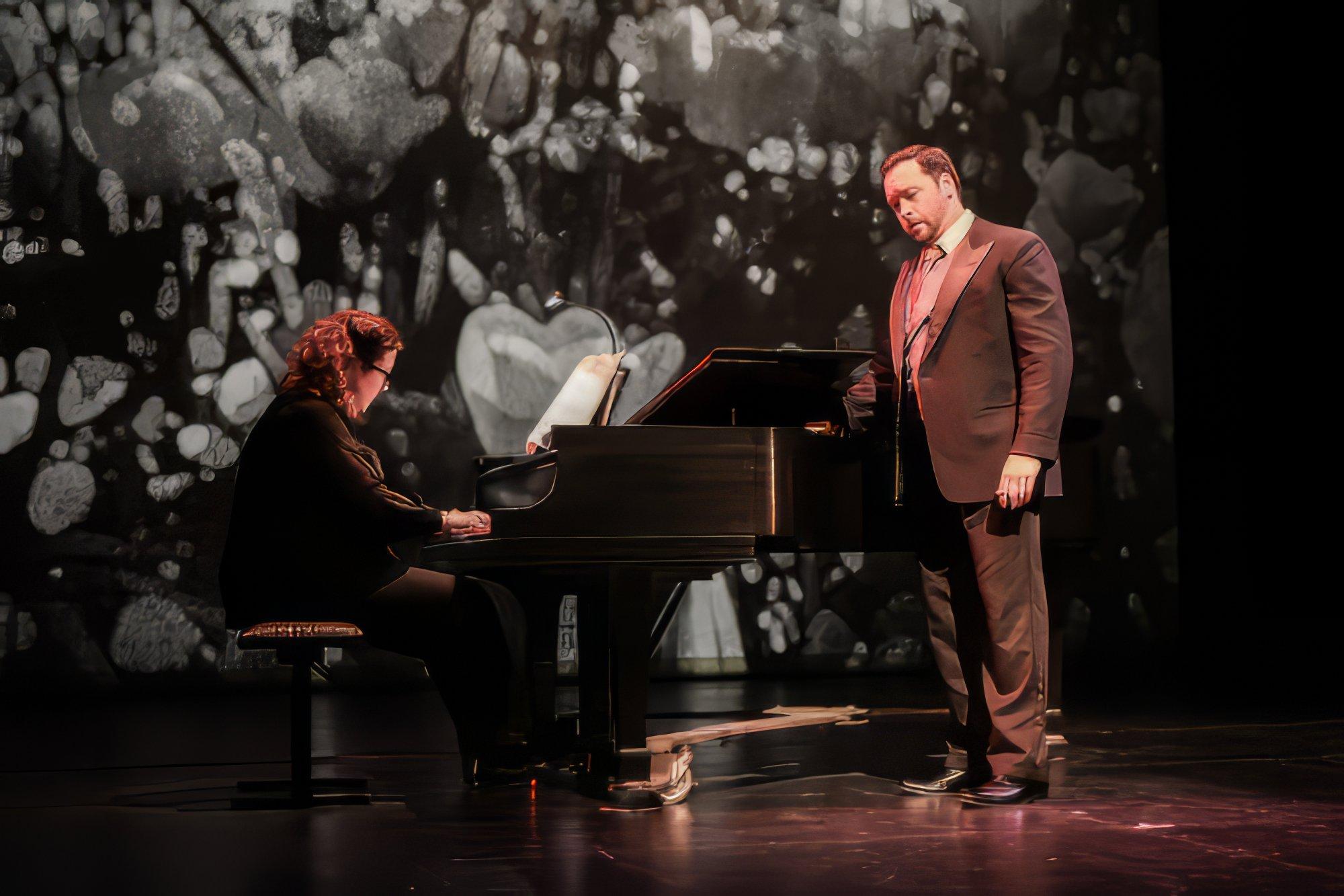 a man in a brown suit sings on stage, accompanied by a pianist behind him