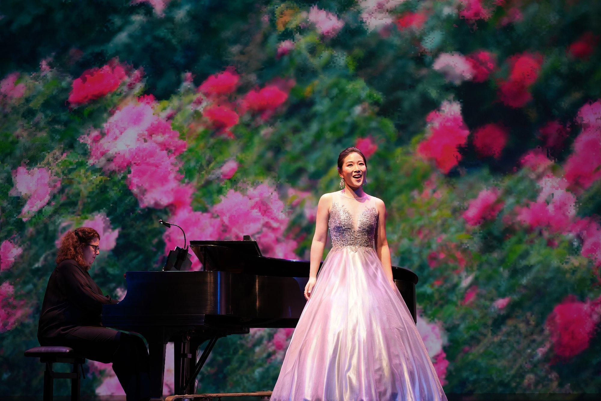 Production Still: A woman stands on stage in formal pink gown with a pianist behind her. A garden of pink flowers is projected on the stage behind her.