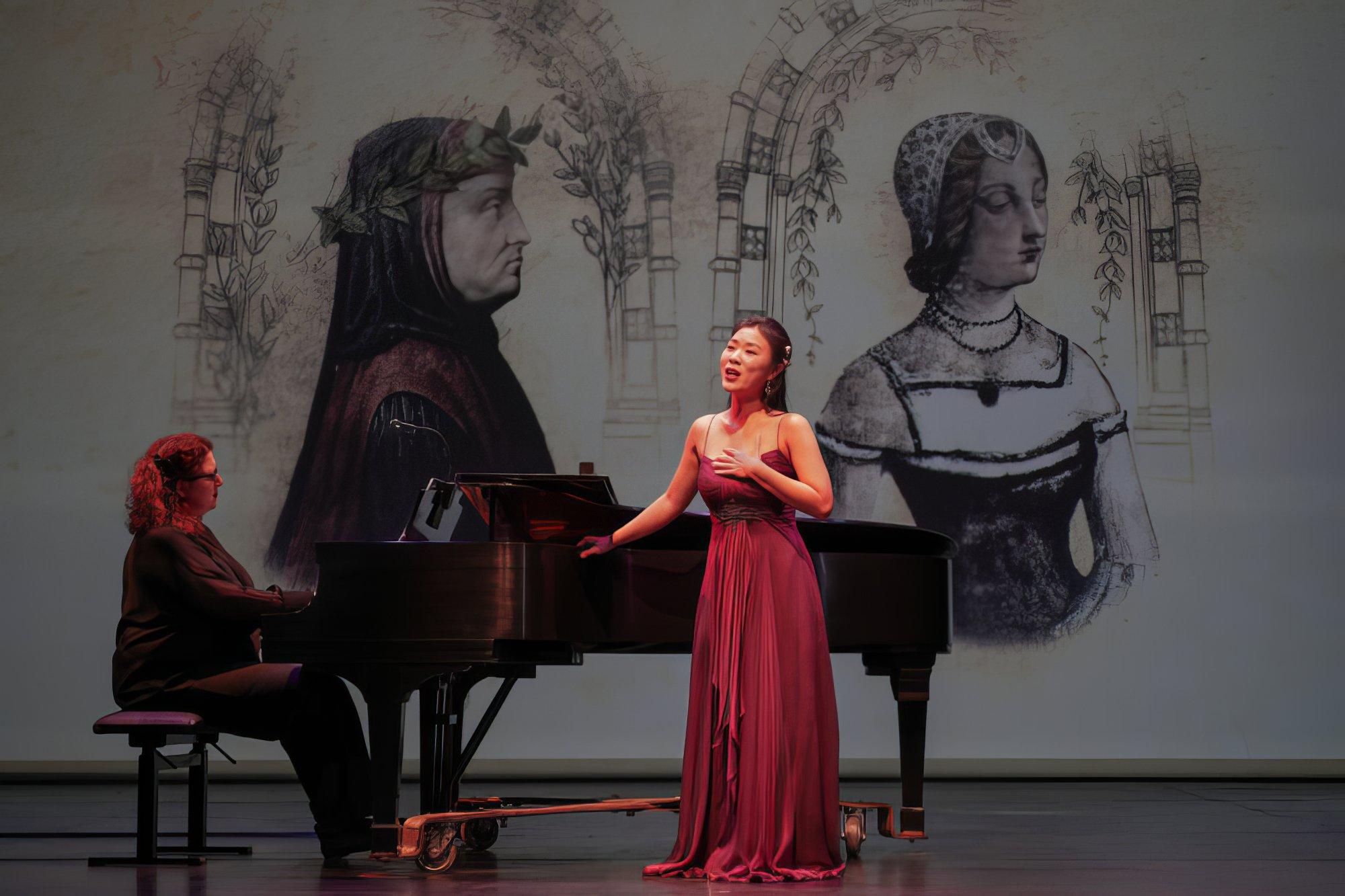 A woman in a red gown sings on stage with a pianist behind here. Sketches of Grecco-Roman busts are projected in the background.