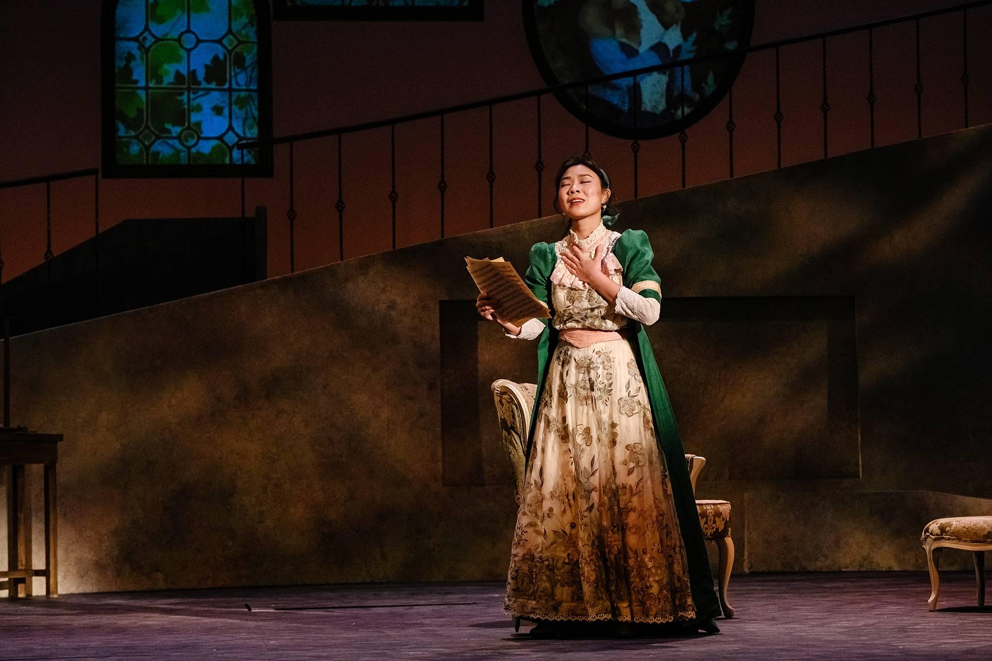 A woman in a beautiful green victorian gown stands alone on stage, holding a letter, and singing solemnly. 