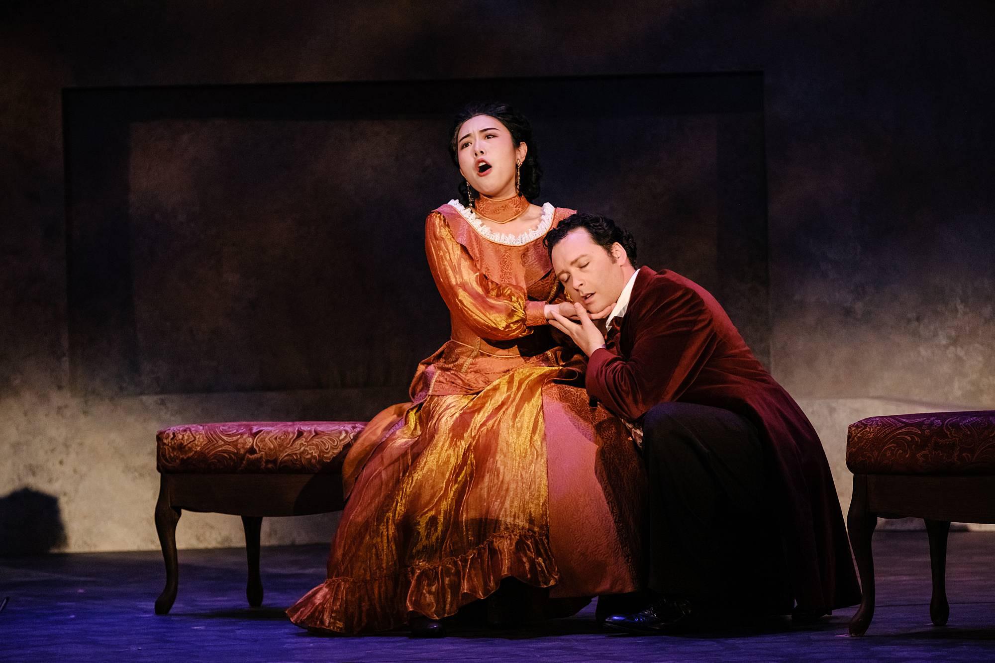 A woma in an ornate gold dress sits on a chaise lounge on a dimly lit stage, singing an aria, while a man in a velvet dinner coat crouches beside her, with his head in her lap.