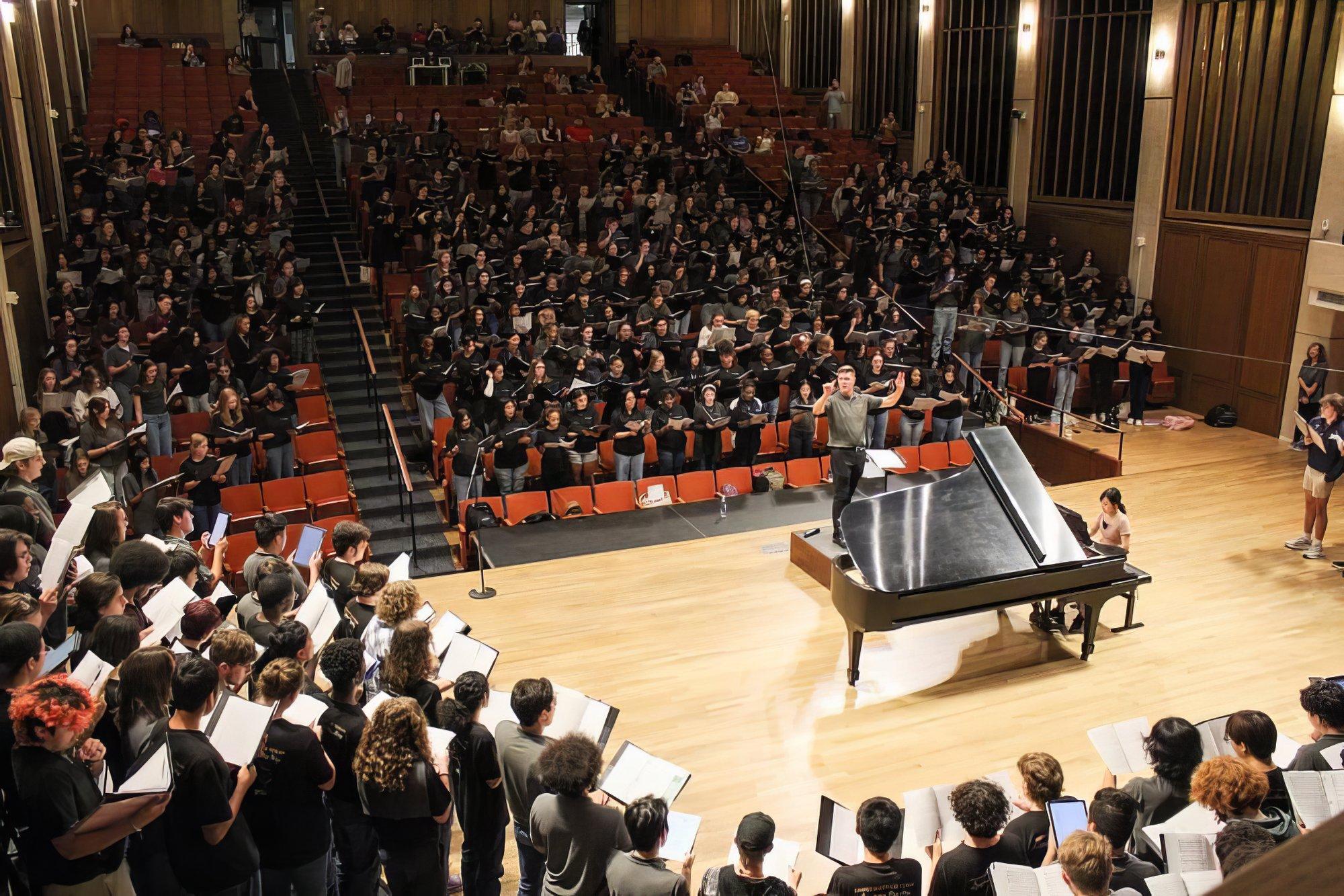 Professor JD Burnett conducts a huge ensemble of choir students on the Bates Hall stage, while more singers fill audience seats behind him, also joining in song.