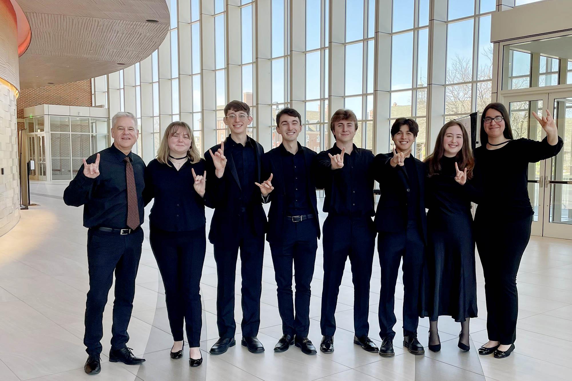 Members of the horn studio stand inside a sleek modern atrium, dressed in black concert dress, they make the hook 'em hand gesture for the camera.