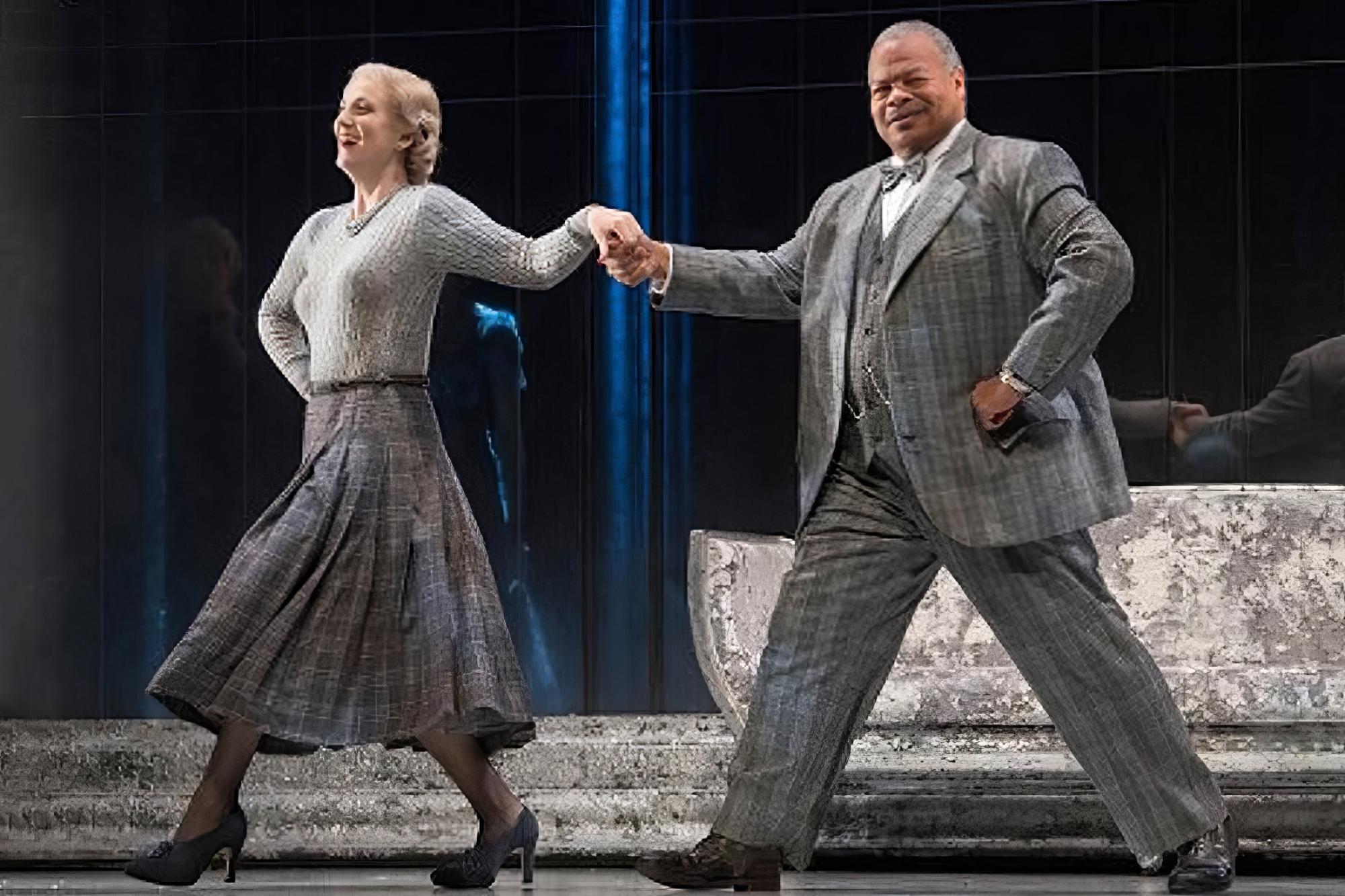 Donnie Ray, dressed in a grey three-piece suit joyfully strides across the stage, escorting a woman as he goes.