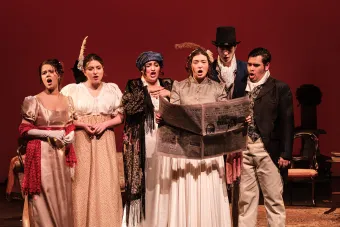 An opera cast huddles around a newspaper looking worried during an opera performance.
