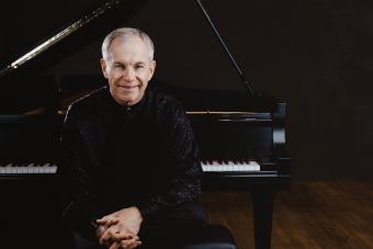 A man with gray hair wearing a black shirt smiles and sits in front of an open piano.