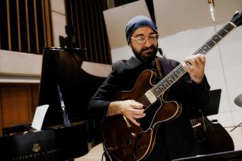 A bearded man wearing a blue beanie and glasses plays an electric bass.