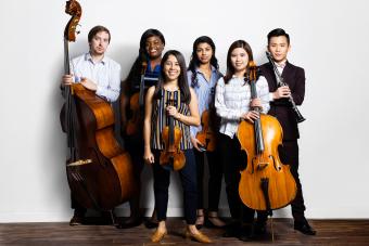 Six students stand together holding string instruments and smiling.