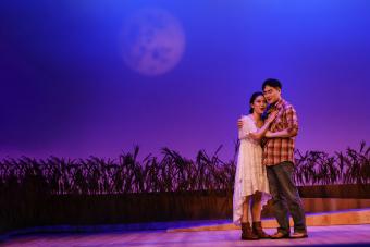 A man and woman embrace under a full moon background on the opera stage