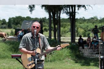 A Man in Pink sunglasses play Electric Guitar and Sings into a microphone on an outdoor stage