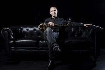Miguel Zenón sits in a dark room on a black sofa with his saxophone across his lap
