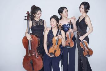 Four asian women hold their string quartet instruments and smile at one another. They are wearing formal concert attire.