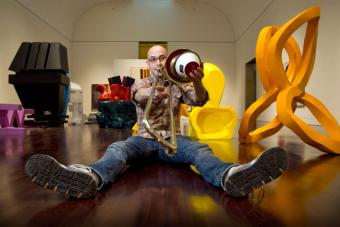 Steven Parker sits on the floor of a modern sculpture gallery, feet stretched in front of him, playing his trombone with a mute