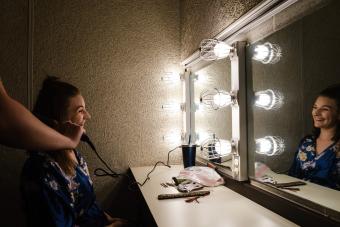 a performer sits, smiling into a mirror lit with globe lights while a hairdresser curls her hair before a show