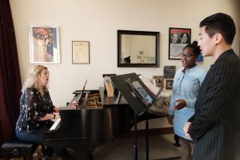 two students sing while their professor accompanies them at the piano in a teaching studio.