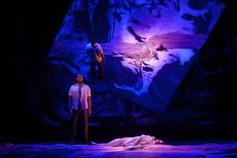 A man towers over a woman collapsed and unconscious on the stage, with a menacing appearance about him. Projected on the background behind him, is a view of the stage from above, creating an eerie, otherworldly mirror-like effect.