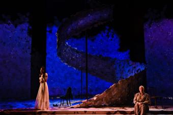 a woman in a flowing white dress sing on an expansive set awash in blue light. Another woman sits on the steps of the set on the opposite side of the stage.  In the middle of the set is a two-story tall spiral made of paper.