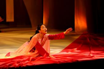 A woman kneels on stage, with an arm outstretched in front of her, surrounded by red fabric, she sings sorrowfully.