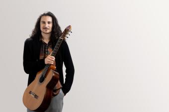 a guitar student holds his guitar across his body at an angle, with one hand in his pocket, looking confident.