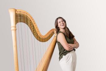 A harp student leans with her back against her harp, arms folded, and smiling.