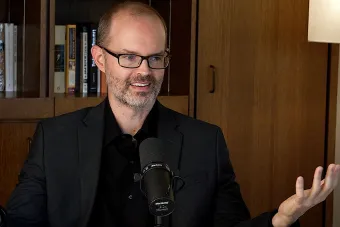 Chris Prosser Talks into a mic with a bookshelf in the background