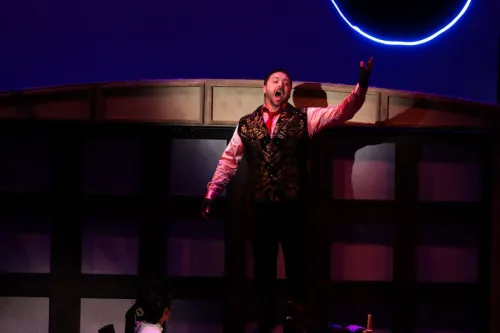 A man stands on stage with arms outstretched below a black moon with a ring of blue neon light around it. Another man sleeps on a mattress on the at his feet. 