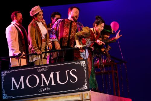 A group of singers stands on a balcony looking angrily at the ground below them. a sign that reads "Momus" is affixed to the balcony in front of them. 