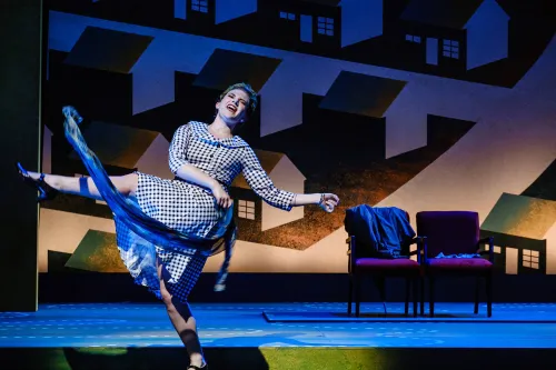 A woman in a plaid mid-century dress kicks her leg into the air as she waves a scarf around during a dance number.