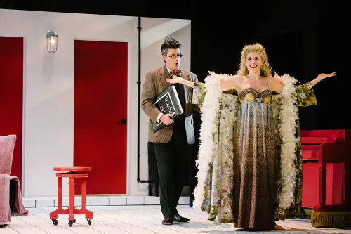 A woman in an ornate night gown and feather boa stands smiling, with her arms outstretched, while a worried conductor holding an opera score looks on behind her.