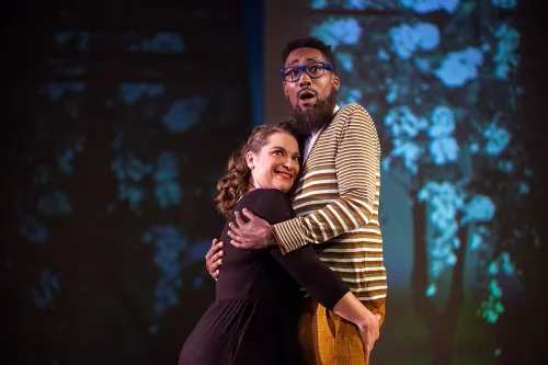 A man and woman embrace each other center stage in front of a forest projected onto the background of the stage.