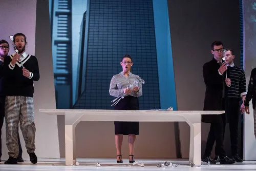 A group of singers in modern business attire stand on the set of a high-rise office building near a large white desk.