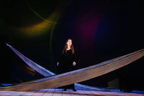 a performer in a black gown stand center stage, long strips of fabric cross-cross in front of her and behind her.