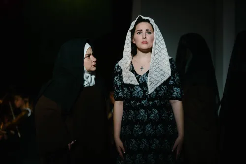 a nun looks at a woman wearing a simple dress and a shawl. The woman looks very frightened.