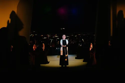 a Priest stands alone in a pool of light on a dark stage, shadows of unseen people are cast on warm orange walls in the background.