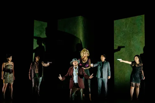 a man in the foreground pleads for his life on his knees, while several performers point guns at him. A woman behind him rests her hands on his shoulders. 