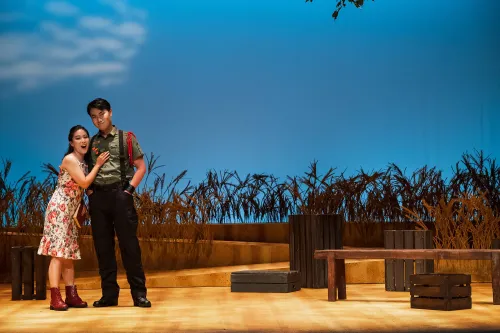 a woman embraces a man on a sun-drenched stage, a field of wheat behind them.