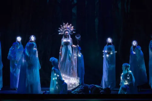 a witch stands on stage in ornate dress surrounded by shadowy figures.