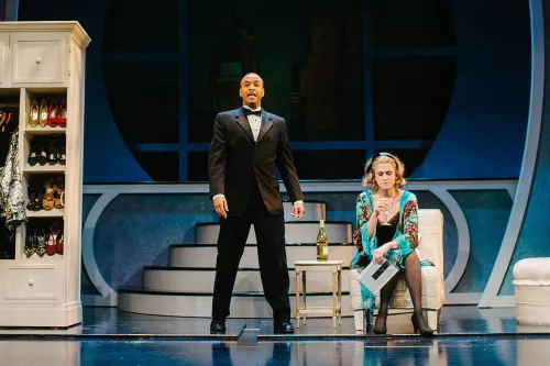 A Man in a tuxedo and a woman in a dressing robe stand near a wardrobe and a night stand.