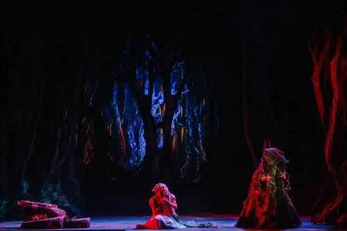 the witch stands hunched over on a wooded set lit by eerie red light while a young girl cowers on her knees, center stage.
