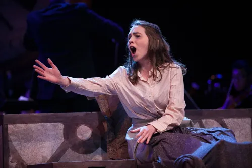 A woman sings as she reaches her arm out to her right, as if she is reaching for someone else. 