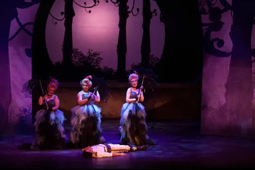 Three women in Elizabethan wigs and dresses stand in an enchanted forest holding parasols, they have stumbled across a man lying unconscious on the ground. 