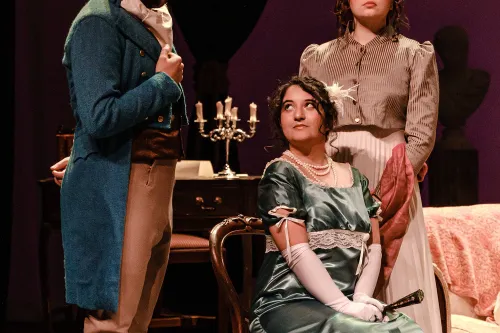 A woman is seated on a chair, flirting innocently with a man standing to her left, dressed in top hat and tails.  An attendant stands behind her, looking unapproving.