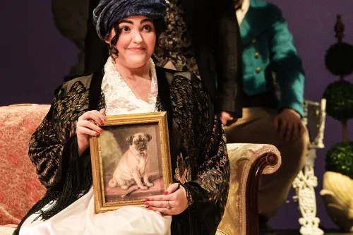 A matronly woman sits tall on a couch proudly displaying a framed portrait of her pug. 