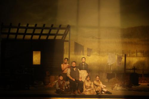 a group of singers clad in pioneer costume sits solemnly on a stage drenched in yellow light in front of a ramshackle living structure and a clothesline holding a few dreary garments.